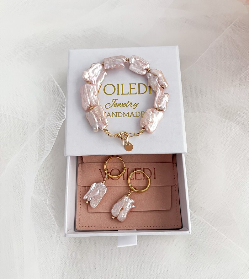 Pu&ograve; includere: Una scatola bianca con un interno rosa che contiene un braccialetto e orecchini di perle. Il braccialetto e gli orecchini sono in oro e hanno un design a forma di perla. La scatola ha il testo "VOILEDI Jewelry HANDMADE" sul coperchio.