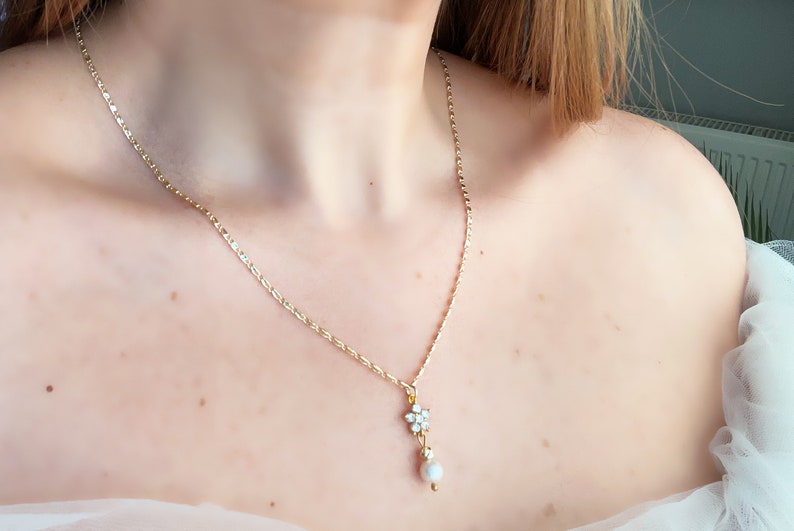 May include: A gold chain necklace with a small flower pendant and a pearl drop. The pendant is made of clear rhinestones.