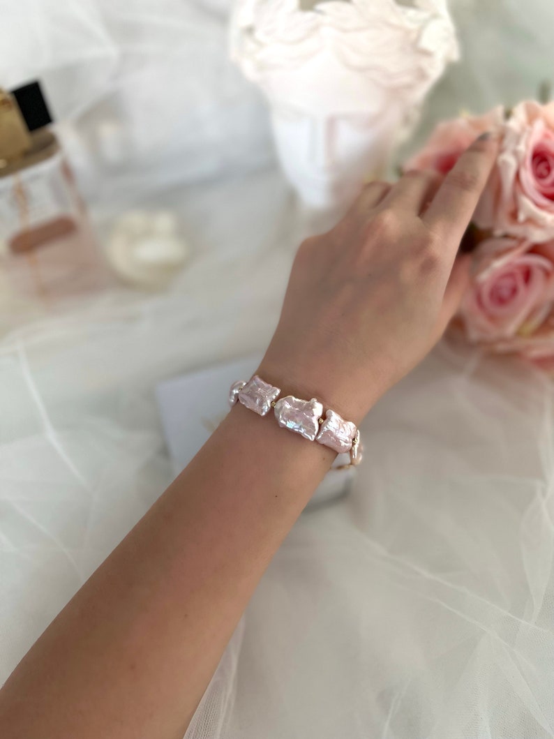 Pu&ograve; includere: Un braccialetto fatto di perle di forma irregolare, con una tonalit&agrave; rosa chiaro. Il braccialetto &egrave; al polso di una persona.
