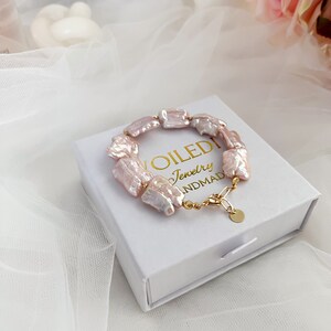 Pu&ograve; includere: Un braccialetto di perle d'acqua dolce rosa con una chiusura e una catena dorate. Il braccialetto &egrave; in una scatola bianca con il testo "VOILEDI Jewelry HANDMAD" stampato su di essa.