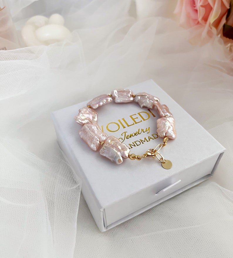 Pu&ograve; includere: Un braccialetto di perle d'acqua dolce rosa con una chiusura e una catena dorate. Il braccialetto &egrave; in una scatola bianca con il testo "VOILEDI Jewelry HANDMAD" stampato su di essa.