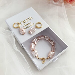 Pu&ograve; includere: Una scatola bianca con il testo "VOILEDI Jewelry HANDMADE" sul coperchio. All'interno della scatola ci sono un paio di orecchini di perle rosa, un braccialetto di perle rosa e due cerchi d'oro.
