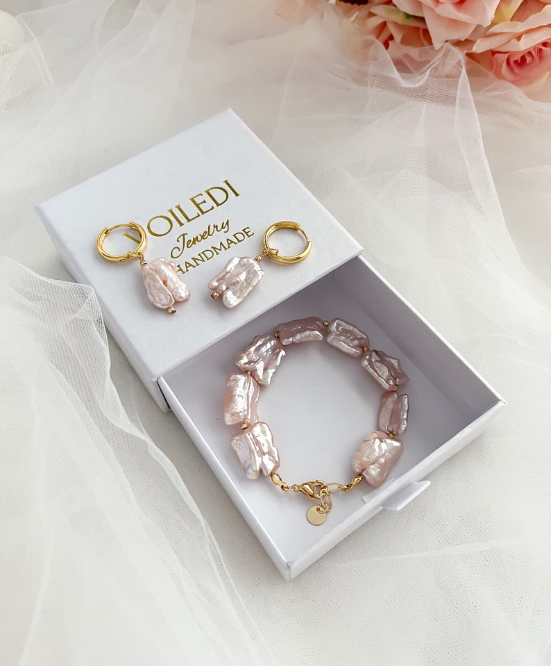 Pu&ograve; includere: Una scatola bianca con il testo "VOILEDI Jewelry HANDMADE" sul coperchio. All'interno della scatola ci sono un paio di orecchini di perle rosa, un braccialetto di perle rosa e due cerchi d'oro.