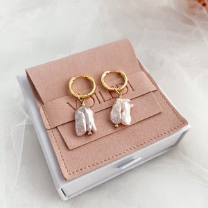 Pu&ograve; includere: Un paio di orecchini a cerchio dorati con un'unica perla barocca pendente da ogni cerchio. Le perle sono di colore rosa e bianco tenue. Gli orecchini sono esposti in una scatola per gioielli in velluto rosa.