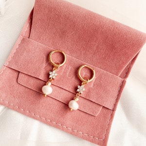May include: A pair of gold hoop earrings with a pearl and a small flower design with clear rhinestones. The earrings are displayed on a pink velvet pouch.