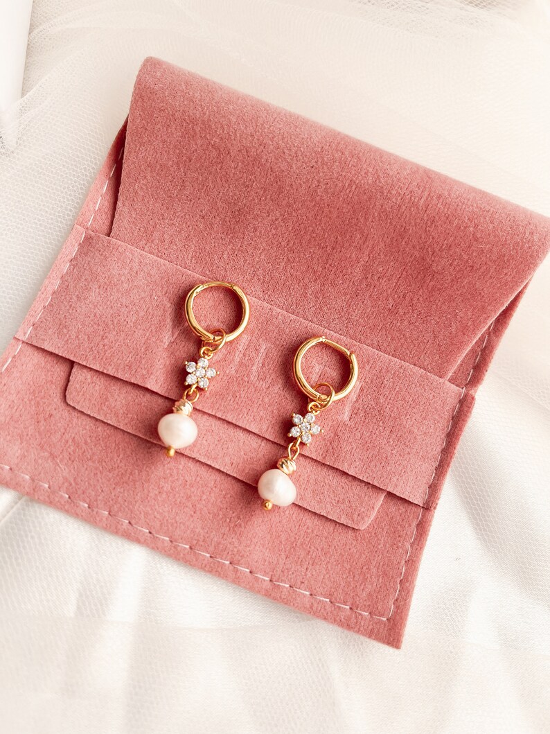 May include: A pair of gold hoop earrings with a pearl and a small flower design with clear rhinestones. The earrings are displayed on a pink velvet pouch.
