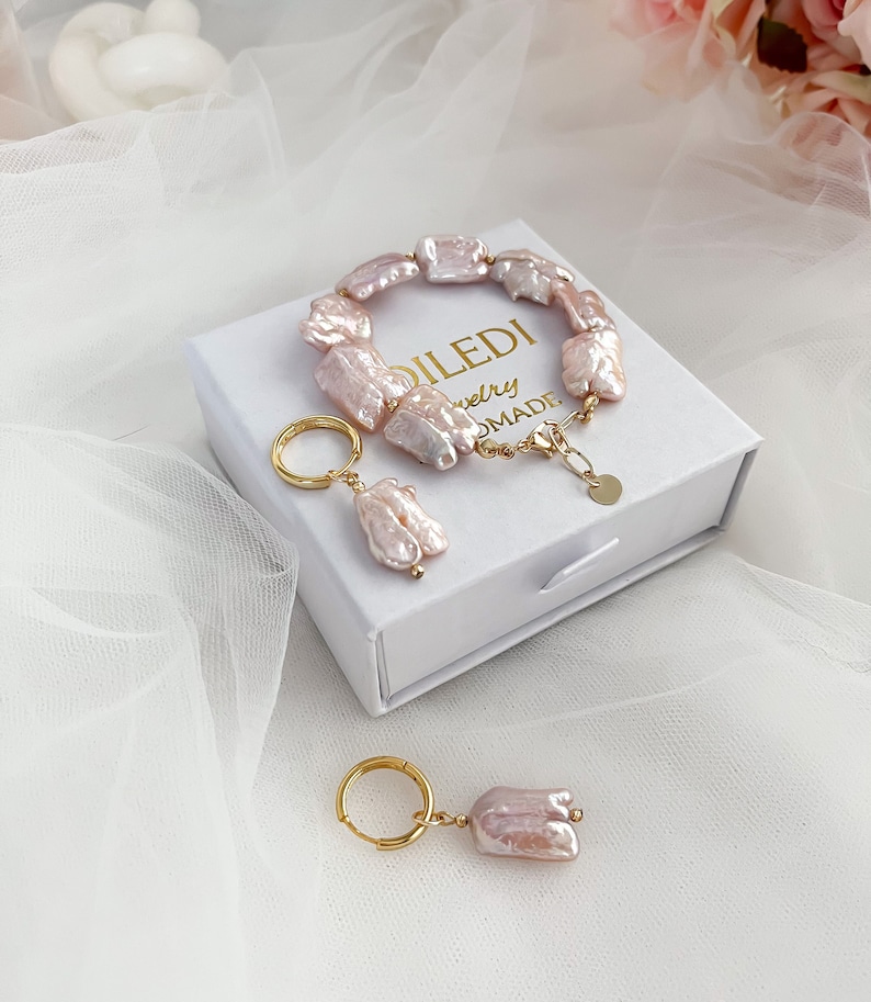 Pu&ograve; includere: Un set di gioielli di perle in una scatola bianca. Il set include un braccialetto e un paio di orecchini. Il braccialetto &egrave; fatto di perle barocche rosa e una catena d'oro. Gli orecchini sono fatti di perle barocche rosa e cerchi d'oro. La scatola &egrave; bianca con lettere dorate che dicono "DILEDI Jewelry Handmade".