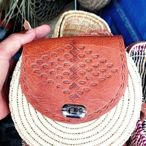 May include: A round, woven straw bag with a brown leather flap and a metal clasp. The bag has a decorative pattern on the leather flap.