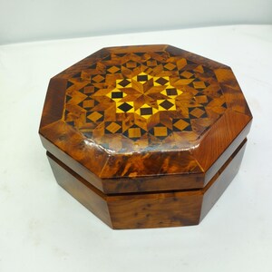 May include: Octagonal wooden box with a geometric inlay pattern on the lid. The inlay is made of different colored woods, including brown, black, and yellow. The box is made of a dark brown wood.