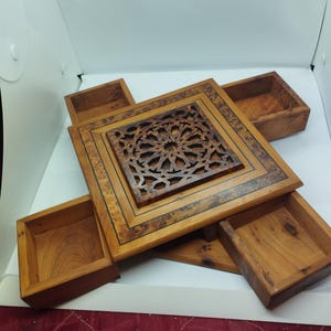 Könnte beinhalten: Holzschmuckkasten mit quadratischer, geschnitzter Oberseite mit geometrischem Design. Die Box hat vier ausziehbare Schubladen und einen dekorativen Rand. Das Holz hat einen warmen, natürlichen Farbton mit dunkleren Akzenten.