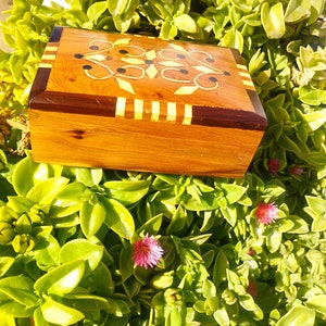 May include: A rectangular wooden box with a hinged lid. The box is made of light brown wood with darker brown accents and decorative inlaid patterns in yellow and black. The box is resting on a bed of green leaves with small pink flowers.