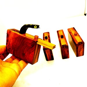 May include: A set of four wooden boxes with a snake-shaped handle on one of the boxes. The boxes are made of dark wood with a reddish hue and have a natural wood grain. The snake handle is black with yellow spots.