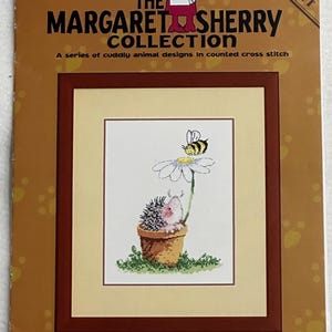 Puede incluir: Un patrón de punto de cruz de la "Colección Margaret Sherry" con una imagen enmarcada de un erizo en una maceta, una margarita y una abeja. El texto "Buzzing By" está en la parte inferior.