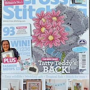 Könnte beinhalten: Titelseite der Zeitschrift 'The World of Cross Stitch' mit einem grauen Tatty Teddy-Bären, der rosa Blumen hält. Das Cover enthält den Text "Britain's No.1" und "Tatty Teddy's BACK!" mit Kreuzstichmustern.