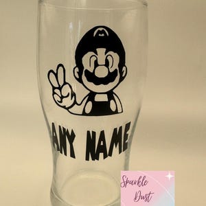 May include: A clear glass featuring a black Mario graphic making a peace sign. The words "ANY NAME" are printed in black below the graphic. The bottom right corner has a logo that says "Sparkle Dust Company".
