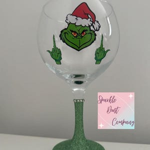May include: A clear wine glass with a green glitter stem and base. The glass features a cartoon Grinch design wearing a Santa hat and giving the middle finger with both hands. The words "Sparkle Dust Company" are on a pink and white square.