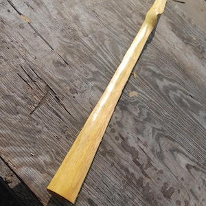 May include: A long, slender piece of light-colored wood, possibly a craft supply or component. The wood has a natural, textured grain and a slightly curved shape. The piece is lying on a weathered wooden surface.