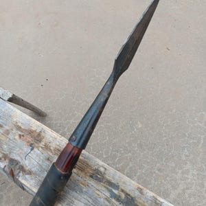 May include: A spear with a long, dark metal blade and a wooden shaft wrapped in black material. The spearhead is sharp and pointed. The shaft has a dark wooden section near the blade and a black grip.