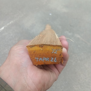 May include: A piece of unfinished lumber, held in a hand, with visible wood grain and a rough texture. The end of the wood is marked with the number "72" and the date "9 APR 26".