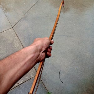May include: A wooden bow with a brown finish, held in a hand. The bow has a curved design with a string attached. The bow is made of wood and has a natural, rustic appearance. The bow is a traditional archery tool.