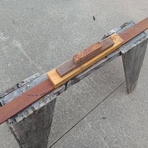 May include: A woodworking scene featuring various wood pieces in different shades of brown and tan. A long, reddish-brown plank rests on a weathered wooden sawhorse, with smaller blocks of wood stacked on top, showcasing the materials used in carpentry.