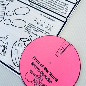 May include: A pink paper wheel with the words "Fruit of the Spirit Secret Decoder" printed on it. The wheel has a small red apple, a bunch of grapes, and a banana drawn on it. The wheel is divided into sections with different fruits and vegetables drawn on each section. The text "Person of Interest" is written on a white sheet of paper with a strawberry illustration.