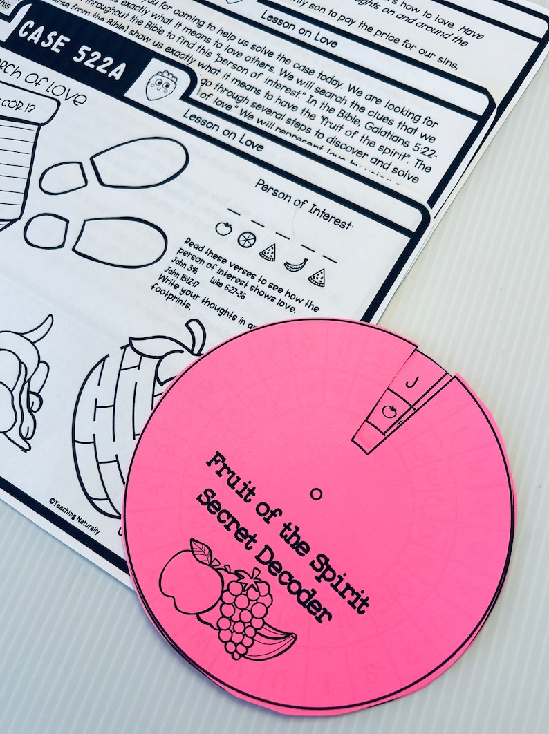 May include: A pink paper wheel with the words "Fruit of the Spirit Secret Decoder" printed on it. The wheel has a small red apple, a bunch of grapes, and a banana drawn on it. The wheel is divided into sections with different fruits and vegetables drawn on each section. The text "Person of Interest" is written on a white sheet of paper with a strawberry illustration.
