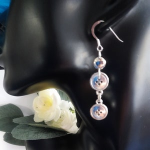 May include: Silver dangle earrings featuring three silver buttons. The buttons have a raised design and are linked by a silver chain.