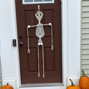 May include: A white string skeleton decoration hangs on a brown door with a white frame. There are pumpkins and gourds on the ground in front of the door.