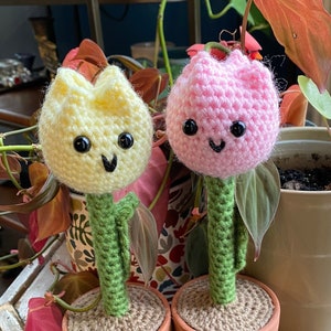 May include: Two crocheted flower pot decorations, one yellow and one pink, with smiling faces and green stems. The pots are brown terracotta.