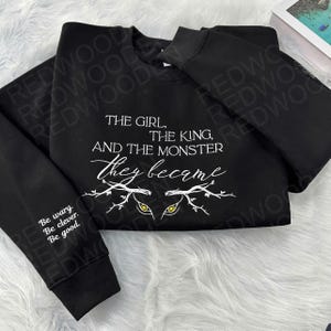 Peut inclure: Sweat-shirt noir avec l'inscription blanche : "THE GIRL, THE KING, AND THE MONSTER they became." Le motif comprend des yeux stylis&eacute;s et des branches. La manche gauche porte l'inscription : "Be wary. Be clever. Be good."
