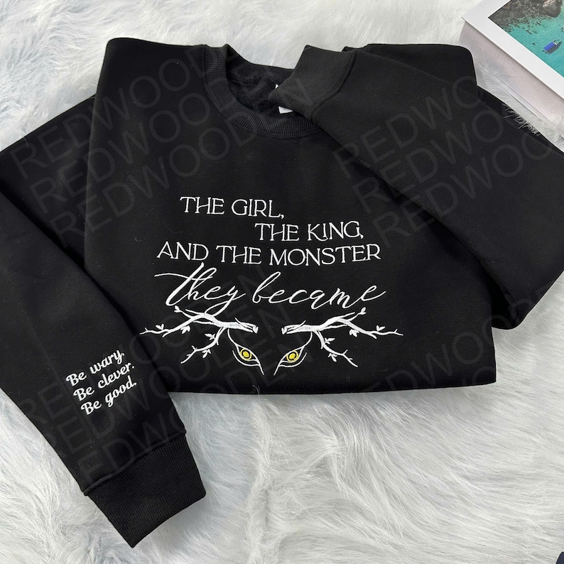 Peut inclure: Sweat-shirt noir avec l'inscription blanche : "THE GIRL, THE KING, AND THE MONSTER they became." Le motif comprend des yeux stylis&eacute;s et des branches. La manche gauche porte l'inscription : "Be wary. Be clever. Be good."