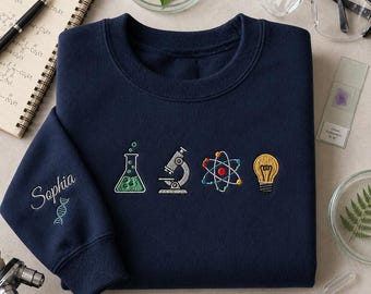May include: A navy blue sweatshirt embroidered with science symbols: a beaker, microscope, atom, and lightbulb. The left sleeve has the name "Sophia" and a DNA strand. Lab equipment and a notebook are in the background.