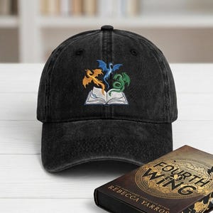 May include: Black denim baseball cap featuring an embroidered design of three dragons in orange, blue, and green above an open book. The cap is a classic six-panel design with a curved brim.
