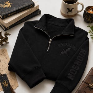 May include: Black quarter-zip sweatshirt with the word "WINGLEADER" and a dragon graphic embroidered on the chest. The sleeve features the name "RIDORSON". The sweatshirt is displayed with dragon-themed books, a mug, and a candle.
