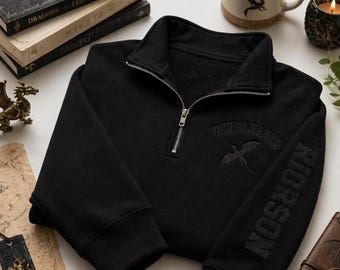 May include: A black quarter-zip sweatshirt featuring a silver metal zip. The chest displays an embroidered dragon silhouette and the text WINGLEADER, while the left sleeve features the embroidered text RIORSON in a matching black thread.