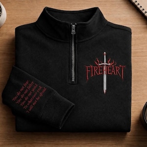 May include: Black quarter-zip sweatshirt featuring a red and silver "FIREHEART" design with a sword. The left sleeve has embroidered text. The sweatshirt is on a wooden surface with stationery and a coffee cup.