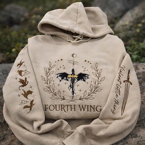 Könnte beinhalten: Beigefarbener Kapuzenpullover mit Kapuze und langen Ärmeln. Die Vorderseite zeigt eine detaillierte Grafik mit einem Schwert, Flügeln und dem Text "FOURTH WING". Der linke Ärmel hat ein Design von fliegenden Kreaturen, und der rechte Ärmel den Text "Violent Little Thing" und ein Schwert.