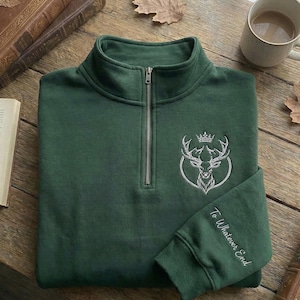 Könnte beinhalten: Ein dunkelgrünes Sweatshirt mit Viertelreißverschluss und silbernem Reißverschluss. Das Sweatshirt hat ein gesticktes Hirschdesign mit Krone und den Worten "To Whatever End" auf dem Ärmel. Das Sweatshirt liegt auf einer Holzoberfläche.