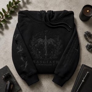 Peut inclure: Un sweat à capuche noir avec une broderie sombre représentant un dragon entouré d'une couronne de feuilles et de phases lunaires. Le texte BASGIATH WAR COLLEGE est centré en dessous. Les manches présentent des silhouettes de dragons et d'épées.