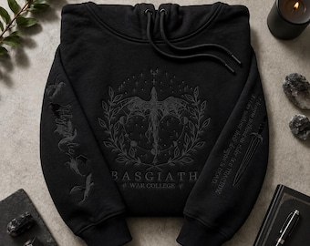 May include: A black hooded sweatshirt featuring a dark gray embroidered dragon design surrounded by a circular wreath and moon phases. The text BASGIATH WAR COLLEGE is centered below. The sleeves feature dragon silhouettes and sword graphics.