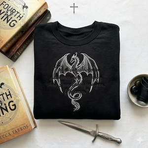 Puede incluir: Sudadera negra con un dise&ntilde;o de drag&oacute;n blanco y el texto "In existence you could find that I wouldn't find you. Violence." La sudadera est&aacute; junto a libros y una daga decorativa.