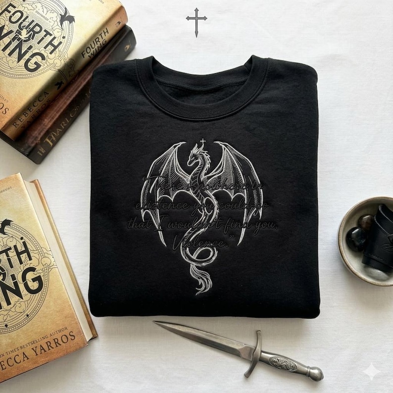 Puede incluir: Sudadera negra con un dise&ntilde;o de drag&oacute;n blanco y el texto "In existence you could find that I wouldn't find you. Violence." La sudadera est&aacute; junto a libros y una daga decorativa.