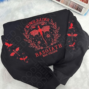 Könnte beinhalten: Schwarzes Sweatshirt mit roter Stickerei. Das Design zeigt den Text "WING LEADER" und "BASGIATH WAR COLLEGE" mit einem Drachen und Sternen. Drachen- und Sternmotive befinden sich auch auf den Ärmeln.