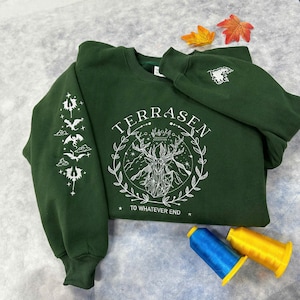 May include: A dark green sweatshirt with a white graphic design featuring a stag with antlers, a crown, and the text "TERRASEN TO WHATEVER END". The graphic is surrounded by a wreath of leaves. The sweatshirt has a white graphic design on the sleeve featuring a series of stylized dragons.