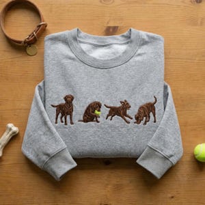 May include: A heather gray crewneck sweatshirt featuring an embroidered design of four brown Labrador retrievers in various poses, including one sitting, one playing with a bright yellow tennis ball, one running, and one sniffing the ground.