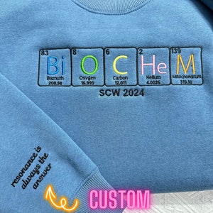 Könnte beinhalten: Ein hellblauer Pullover mit dem Wort "BIOCHEM", das mit den chemischen Symbolen für Bismuth, Sauerstoff, Kohlenstoff, Helium und Mitochondrium ausgeschrieben ist. Der Text "SCW 2024" befindet sich unter den Symbolen. Der Ärmel des Pullovers hat den Text "resonance is always the answer" aufgestickt.