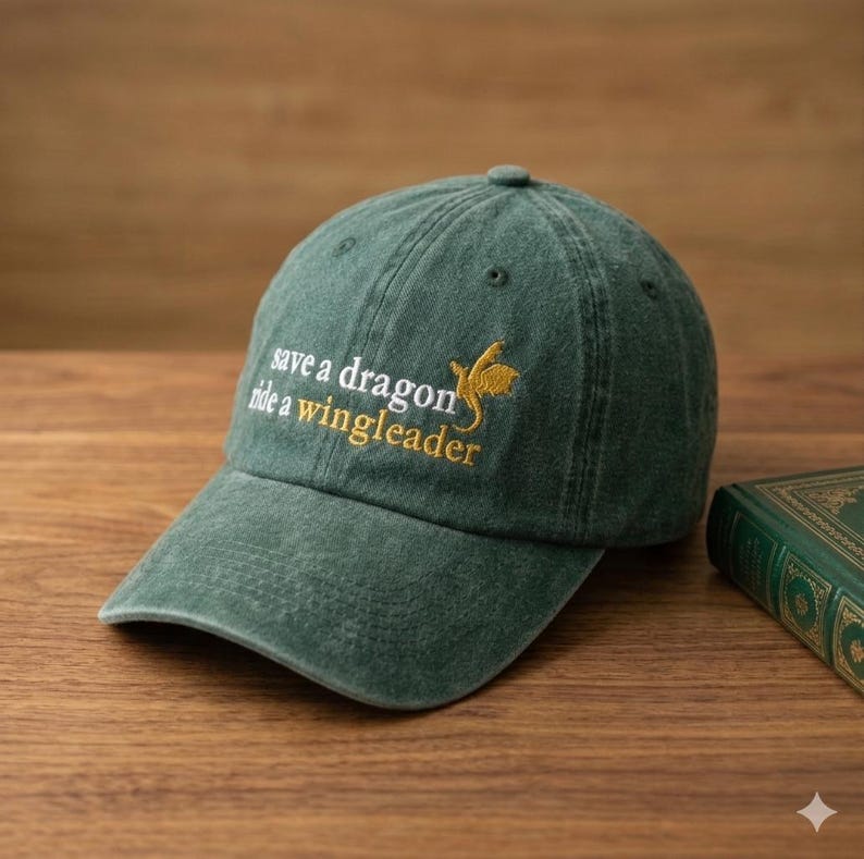 May include: Green baseball cap with the embroidered text "save a dragon ride a wingleader" in white and gold. The cap is made of a soft, worn material and has a curved brim. A book is visible in the background.
