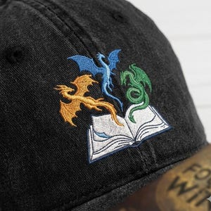 May include: Black baseball cap with an embroidered design of three dragons in gold, blue, and green above an open book. The cap is made of a textured fabric and has a curved brim.
