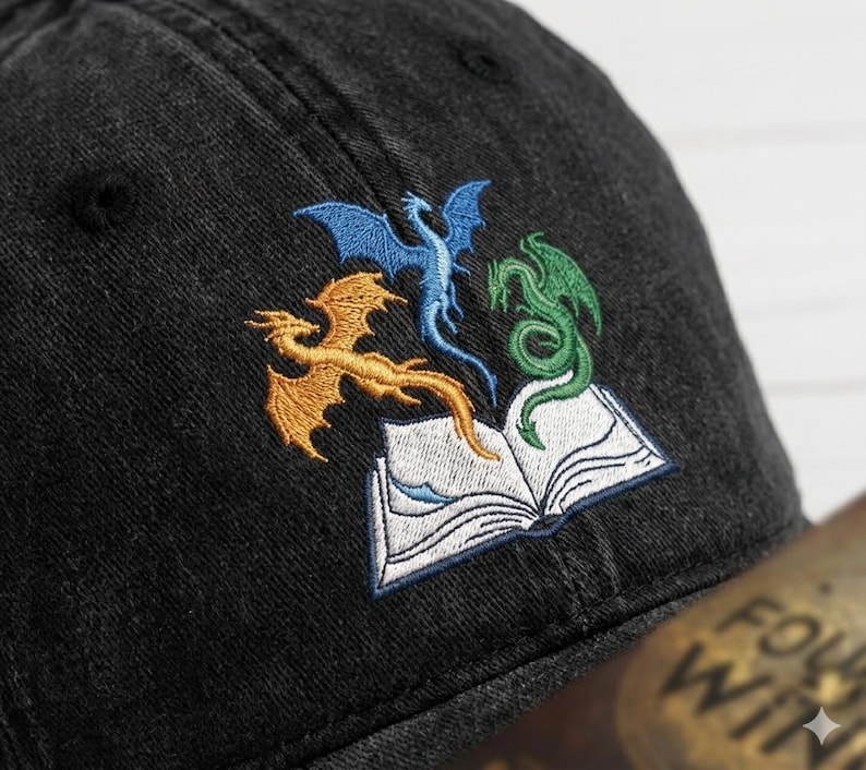 May include: Black baseball cap with an embroidered design of three dragons in gold, blue, and green above an open book. The cap is made of a textured fabric and has a curved brim.